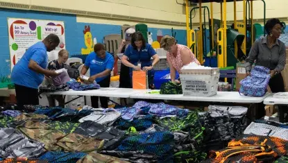 A group of volunteers stand behind a large pile of backpacks, organizing school supplies.