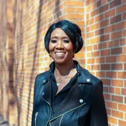 Photo of a smiling Black woman posing in front of a brick wall