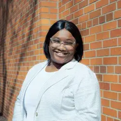 Photo of a smiling Black woman with clear glasses posing in front of a brick wall