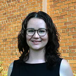 Photo of a smiling white woman with glasses posing in front of a brick wall